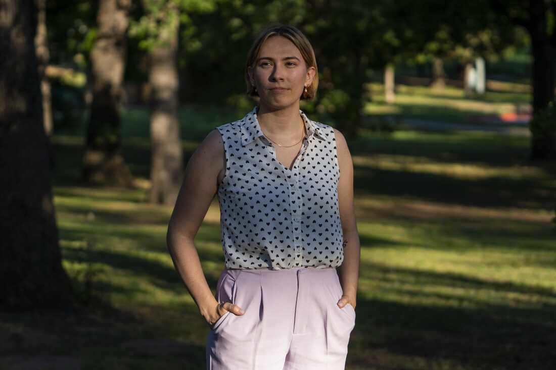 Caitlyn Mai stands in a park near her home in Bethany, Okla., on May 18, 2024. She is wearing light-colored pants and a white sleeveless blouse with small dark hearts.
